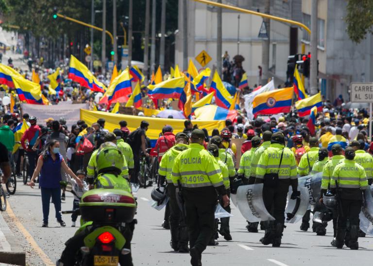 Riot police walk towards protestors holding multiple Colombian flags.
