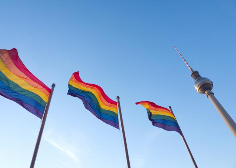 Three Pride flags waving in the wind next to a silver gleaming tower against a big blue sky.