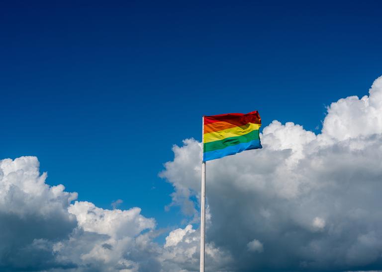 Un drapeau de la Fierté LGBTQ flotte au vent sur un fond de ciel bleu et de nuages majestueux.