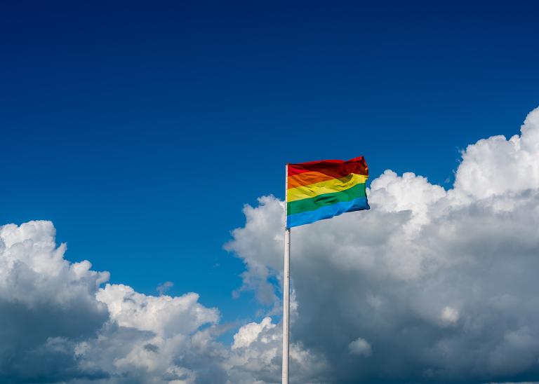 LGBTQ Pride flag flapping in the wind set against a big blue sky with picturesque fluffy white clouds.