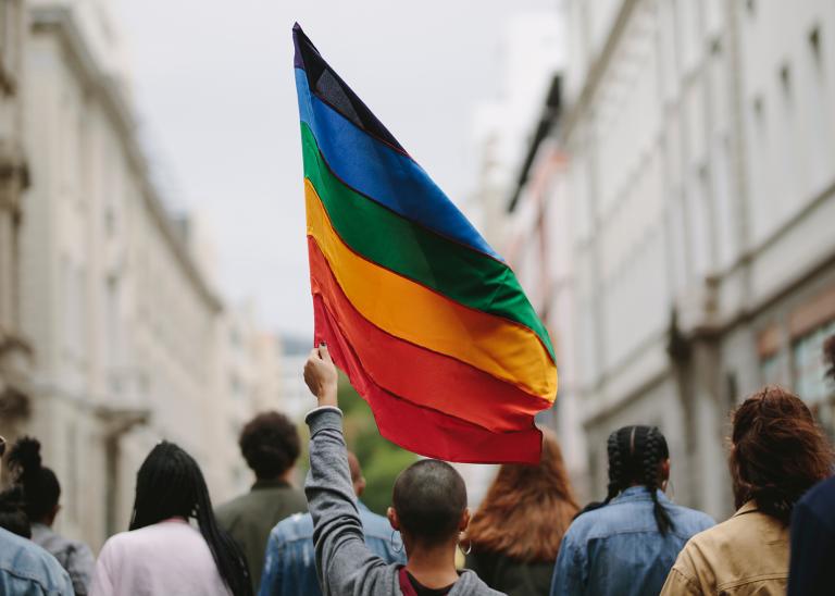 a single LGBTQ pride flag carried by a person walking in a parade, view is of the backs of the people walking along a city street framed by blurred tall buildings on either side