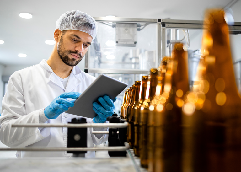 A brewery employee in protective clothing looks at a clipboard with beer bottles in front of him 