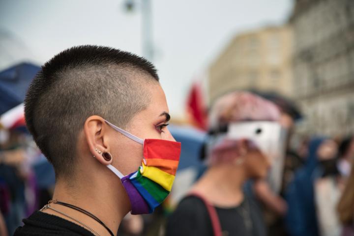 A side profile of a person wearing a Pride flag face mask with a blurred group of people behind them.